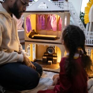 A father and daughter enjoy a playful moment with a dollhouse, featuring a blurred background.