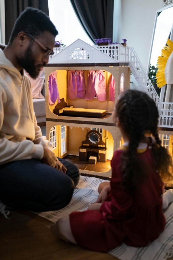 A father and daughter enjoy a playful moment with a dollhouse, featuring a blurred background.