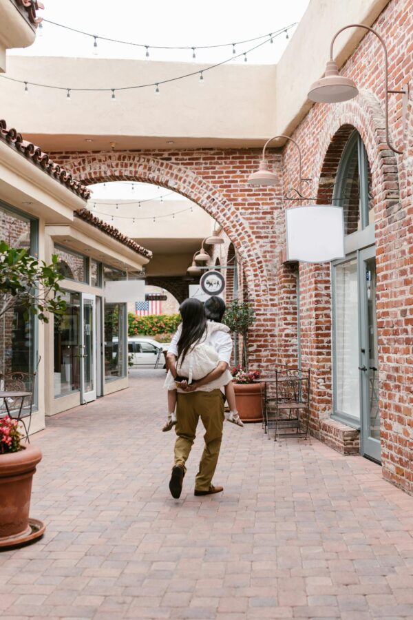 A father gives his daughter a piggyback ride through a charming brick alleyway, embodying family bonding.